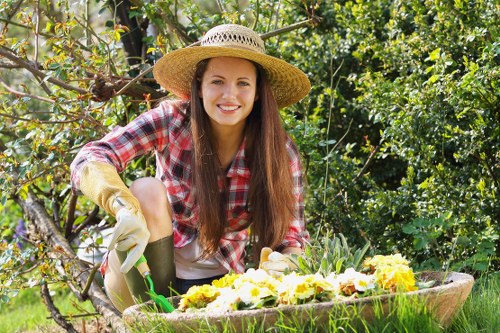 A young woman with long brown hair, wearing a straw sun hat, a red and white checkered shirt, and gardening gloves, is kneeling in a lush garden. She is smiling at the camera while tending to a flower bed filled with blooming yellow and white flowers. The garden features a neatly maintained grassy lawn, surrounded by shrubs and small trees, with a wooden garden border visible in the background. Sunlight filters through the leafy canopy, casting a natural glow over the vibrant greenery and floral display. The scene exudes a healthy, well-kept outdoor space suitable for gardening and landscaping services in Croydon, with the garden showing signs of regular care and attention to detail typical of professional garden maintenance by Gardener Croydon.