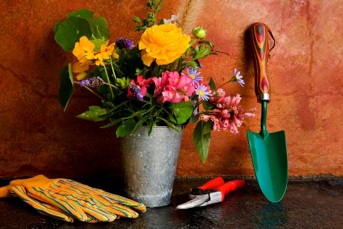 A close-up of a garden scene showing a large, grey concrete flowerpot filled with a mixed arrangement of vibrant flowers including yellow, pink, and purple blooms, along with green foliage. The flowerpot is placed on a dark stone surface against a textured, reddish-brown wall. Nearby, there are gardening tools such as a small green trowel with a multicoloured handle, a pair of pruning shears with red handles, and a pair of yellow and orange-striped gardening gloves. The outdoor environment appears well-lit, suggesting daytime with clear weather, and the setup indicates preparation for gardening activities, reflecting the type of outdoor maintenance services offered by Gardener Croydon in the Croydon area. The scene emphasizes the importance of flower planting and garden tool use in maintaining a beautiful garden space.